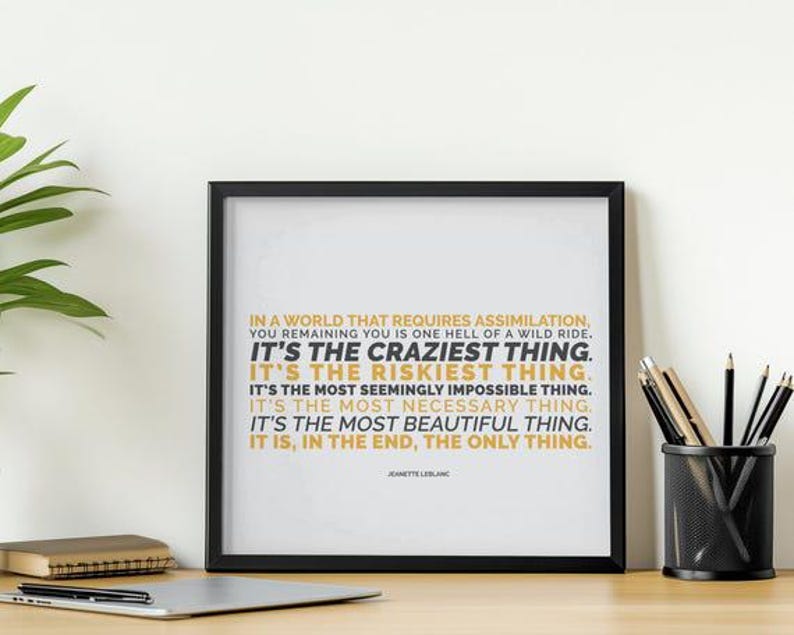 You Remaining You printable shown in a black frame leaning against a white wall on a light wood desk, beside a silver laptop, a black mesh cup of pencils, a small stack of notebooks, and a leafy plant at the edge. Clean, bright daylight. Home office styling for an uplifting quote. A motivational printable for a desk where you do your own work your own way, with the line It is, in the end, the only thing in gold capitals.
