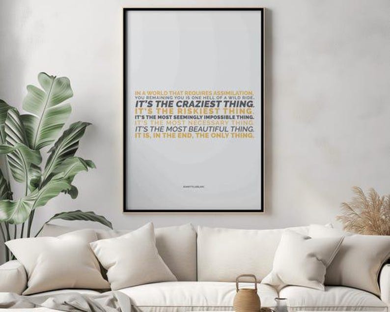 You Remaining You printable shown in portrait layout in a black frame hanging on a soft gray plaster wall above a cream sofa with cream pillows, beside a large banana leaf plant, dried pampas grass, and a small woven basket. The gold and black text sits near the top of the page. Calm living room styling. An independent woman reminder for over the sofa, and a digital download wall art piece you can print to fit your own frame.