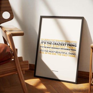 You Remaining You printable shown in a black frame leaning against a white wall on a wood floor, between a wood armchair with a brown leather cushion and a light wood chair, in warm low sun with long shadows. The gold and black lines sit in a short block in the middle of the white page. Mid century styling for a resilience print, a fit for a reading corner or a den where you stop to think about who you are.