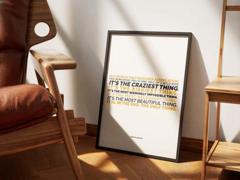 You Remaining You printable shown in a black frame leaning against a white wall on a wood floor, between a wood armchair with a brown leather cushion and a light wood chair, in warm low sun with long shadows. The gold and black lines sit in a short block in the middle of the white page. Mid century styling for a resilience print, a fit for a reading corner or a den where you stop to think about who you are.