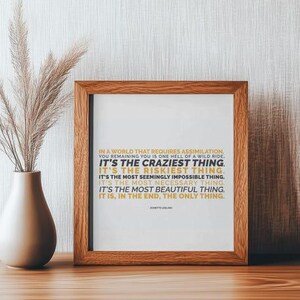 You Remaining You printable shown in a warm brown wood frame standing on a wood shelf against a textured white wall, beside a cream ceramic vase of dried pampas grass. Soft side light and a gentle shadow. Simple neutral decor styling that lets the mustard gold and black text stand out. A thoughtful personal growth gift for a friend learning to stay herself, and an easy piece to print at home or at a local print shop.