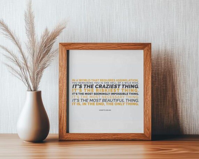 You Remaining You printable shown in a warm brown wood frame standing on a wood shelf against a textured white wall, beside a cream ceramic vase of dried pampas grass. Soft side light and a gentle shadow. Simple neutral decor styling that lets the mustard gold and black text stand out. A thoughtful personal growth gift for a friend learning to stay herself, and an easy piece to print at home or at a local print shop.