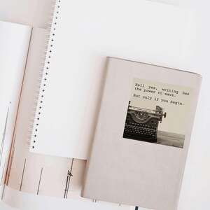Beige notebook decorated with a sticker reading “Hell yes, writing has the power to save. But only if you begin.” Featuring a vintage typewriter, this Jeanette LeBlanc quote inspires creative courage and writing encouragement.