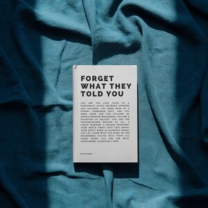 May include: A book titled "FORGET WHAT THEY TOLD YOU" rests on a teal fabric. The book's cover is white with black text. The text on the cover includes a poem about self-discovery and freedom. The fabric has soft folds and shadows.