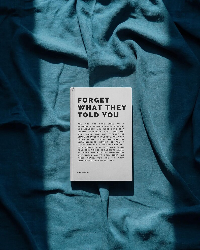 May include: A book titled "FORGET WHAT THEY TOLD YOU" rests on a teal fabric. The book's cover is white with black text. The text on the cover includes a poem about self-discovery and freedom. The fabric has soft folds and shadows.