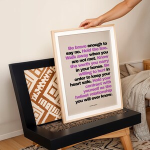 A hand sets the purple and black boundary print, in a light wood frame with a white border, onto a black wood chair with a brown and white patterned pillow, beside a bed with a knit throw and a plant on a side table. Playful, layered styling with warm earth tones. A divorce print idea, or a recovery gift for someone rebuilding her life on her own terms after a hard relationship.