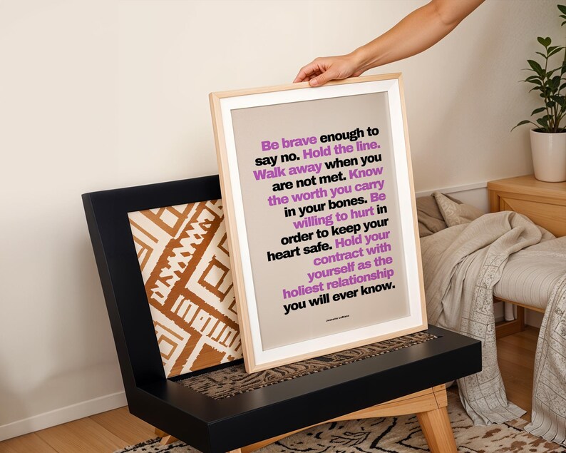 A hand sets the purple and black boundary print, in a light wood frame with a white border, onto a black wood chair with a brown and white patterned pillow, beside a bed with a knit throw and a plant on a side table. Playful, layered styling with warm earth tones. A divorce print idea, or a recovery gift for someone rebuilding her life on her own terms after a hard relationship.
