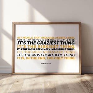 Close view of the You Remaining You printable in a light wood frame, in landscape layout, leaning against a white wall on a sunlit pale wood floor with soft window shadows. Lines of capital text alternate between mustard gold and black, from IN A WORLD THAT REQUIRES ASSIMILATION down to IT IS, IN THE END, THE ONLY THING, with the name Jeanette LeBlanc below. A typography poster about staying true to yourself, sold as printable wall art.