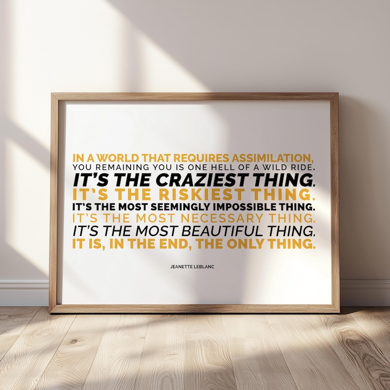 Close view of the You Remaining You printable in a light wood frame, in landscape layout, leaning against a white wall on a sunlit pale wood floor with soft window shadows. Lines of capital text alternate between mustard gold and black, from IN A WORLD THAT REQUIRES ASSIMILATION down to IT IS, IN THE END, THE ONLY THING, with the name Jeanette LeBlanc below. A typography poster about staying true to yourself, sold as printable wall art.