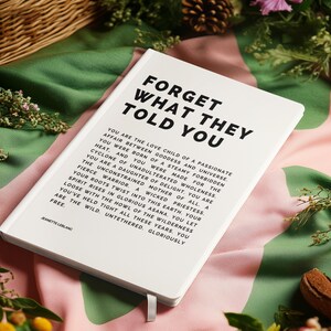 May include: A white hardcover journal with the text "FORGET WHAT THEY TOLD YOU" in bold black letters. The journal has a ribbon bookmark and is surrounded by flowers and greenery. The journal's text is a poem.