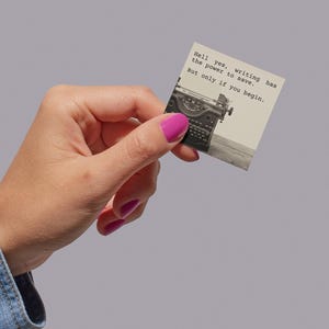 A sticker featuring a vintage typewriter and the words “Hell yes, writing has the power to save. But only if you begin.” Held in a hand, this Jeanette LeBlanc quote is perfect for writing encouragement and inspiration for creatives.
