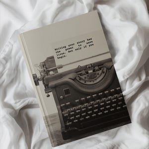 以下が含まれることがあります： 「Writing your story has the power to save lives. But only if you begin.」と書かれた白いノート。ノートが開かれており、背景にはビンテージのタイプライターが見える。