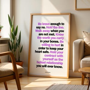 Purple and black boundary print in a light wood frame leaning on a sunny wood floor between a wood armchair with a cream cushion and a cream sofa with a tan pillow, beside a snake plant, a small potted plant on the windowsill, and a round wood side table with books. A striped rug and a white radiator complete the room. Living room styling for a healing quote print in a calm, warm neutral space.
