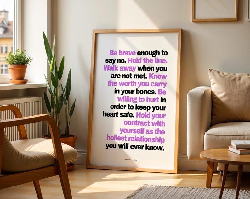 Purple and black boundary print in a light wood frame leaning on a sunny wood floor between a wood armchair with a cream cushion and a cream sofa with a tan pillow, beside a snake plant, a small potted plant on the windowsill, and a round wood side table with books. A striped rug and a white radiator complete the room. Living room styling for a healing quote print in a calm, warm neutral space.