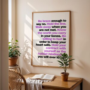 Purple and black boundary print in a light wood frame hanging on a cream wall above a small wood desk with an open book, a white cloth, and a potted plant, beside a rattan chair and a plant on a stand. Soft shadows of leaves fall on the wall. A writing desk idea for a journaling or shadow work space, and a quiet piece of office wall art for a home study.