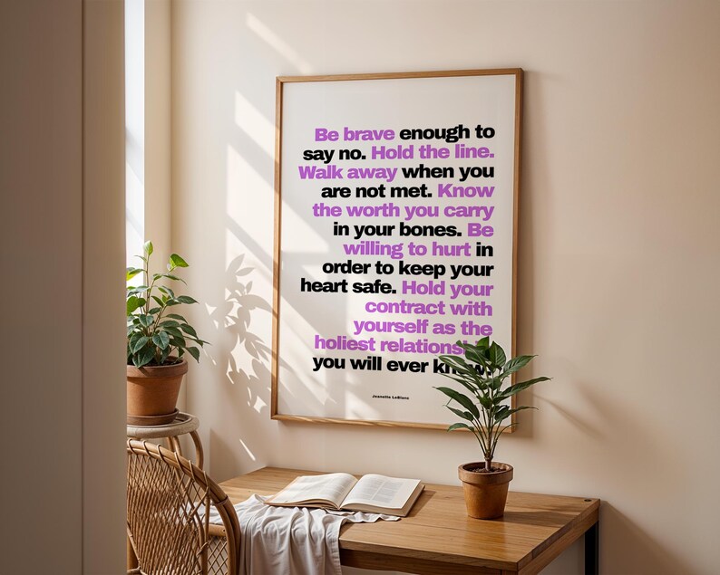 Purple and black boundary print in a light wood frame hanging on a cream wall above a small wood desk with an open book, a white cloth, and a potted plant, beside a rattan chair and a plant on a stand. Soft shadows of leaves fall on the wall. A writing desk idea for a journaling or shadow work space, and a quiet piece of office wall art for a home study.