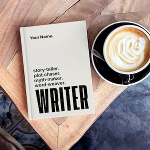 May include: A cream-coloured book with the text "Your Name." and "story-teller. plot-chaser. myth-maker. word-weaver. WRITER" in black. A cup of coffee with latte art sits on a black saucer beside the book.