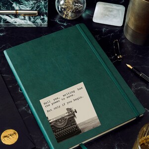 Dark green journal with a sticker reading “Hell yes, writing has the power to save. But only if you begin.” Featuring a vintage typewriter and a Jeanette LeBlanc quote, perfect for writing inspiration and creative motivation.