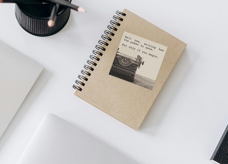A kraft spiral notebook adorned with sticker reading, “Hell yes, writing has the power to save. But only if you begin.” Featuring a vintage typewriter & a Jeanette LeBlanc quote, this design inspires writing encouragement and motivation for writers.
