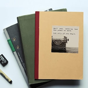 A beige notebook with a sepia-toned typewriter sticker sits atop green and gray books. The sticker reads, “Hell yes, writing has the power to save. But only if you begin.”  Jeanette LeBlanc quote writing inspiration & encouragement for storytellers.