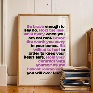 Purple and black boundary print in a light wood frame leaning against white painted wood paneling on a wood floor, beside a tall stack of books with an open book on top and a wood bench with a beige cushion. A white radiator sits at the left. Clean, bright styling for a self love print that pairs well with books and natural wood, from writer Jeanette LeBlanc.