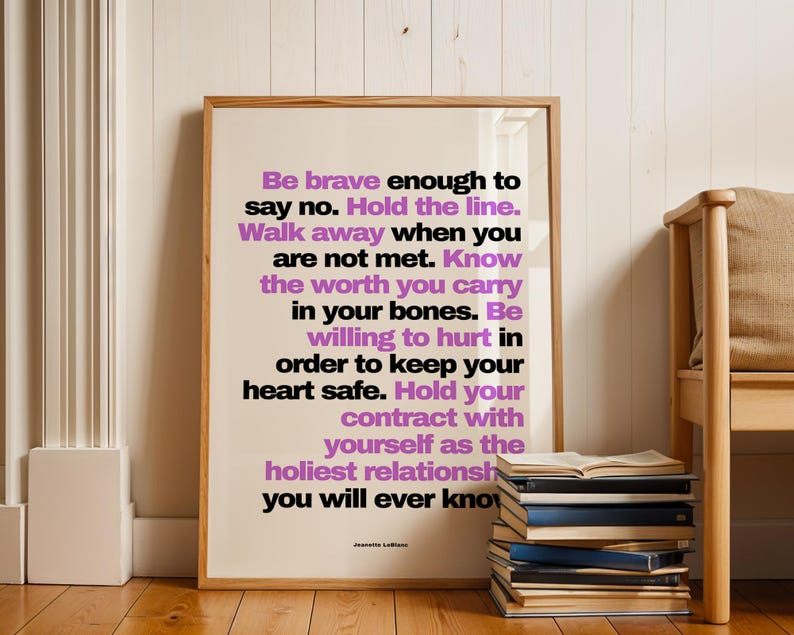 Purple and black boundary print in a light wood frame leaning against white painted wood paneling on a wood floor, beside a tall stack of books with an open book on top and a wood bench with a beige cushion. A white radiator sits at the left. Clean, bright styling for a self love print that pairs well with books and natural wood, from writer Jeanette LeBlanc.