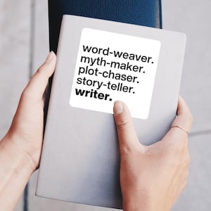 May include: A light gray notebook with the words "word-weaver. myth-maker. plot-chaser. story-teller. writer." printed on a white sticker. The notebook is held in two hands, with a dark blue notebook in the background.