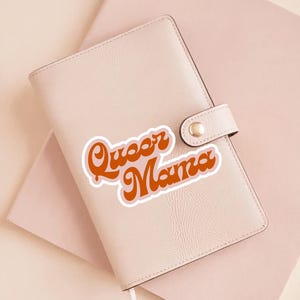 May include: A light pink leather journal with a snap closure, a white ribbon bookmark, and a sticker that reads "Queer Mama" in orange script with a white outline. The journal rests on a light pink surface.