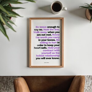 Overhead view of the purple and black boundary print in a light wood frame lying on a white desk, with a cup of black coffee, two potted plants in the corners, and a tan leather chair tucked below on a gray rug. The words Know the worth you carry in your bones stand out in purple. A clean office desk view for a self worth print, and a thoughtful gift for a psychologist opening a new practice.