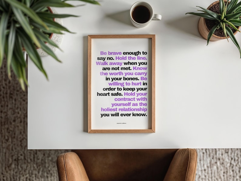 Overhead view of the purple and black boundary print in a light wood frame lying on a white desk, with a cup of black coffee, two potted plants in the corners, and a tan leather chair tucked below on a gray rug. The words Know the worth you carry in your bones stand out in purple. A clean office desk view for a self worth print, and a thoughtful gift for a psychologist opening a new practice.