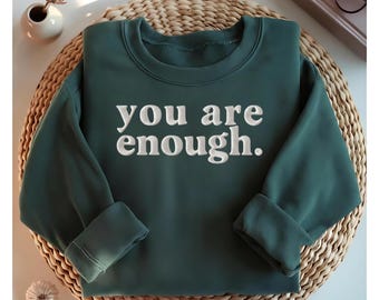 Op de afbeelding: Een donkergroene sweatshirt met de geborduurde tekst "you are enough." De sweatshirt is op een geweven mat gevouwen. De mouwen zijn opgerold. Een kleine witte vaas met een gedroogde bloem staat op de achtergrond.