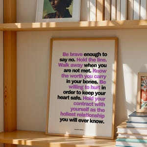 Purple and black boundary print in a light wood frame leaning on a light wood bookshelf, beside a stack of books and a small framed red and blue floral painting, with a framed portrait and a row of books on the shelf above. Warm side light. A bookish wall art moment for a library corner, and a strong woman quote to keep among your favorite books and the authors who taught you to stand firm.