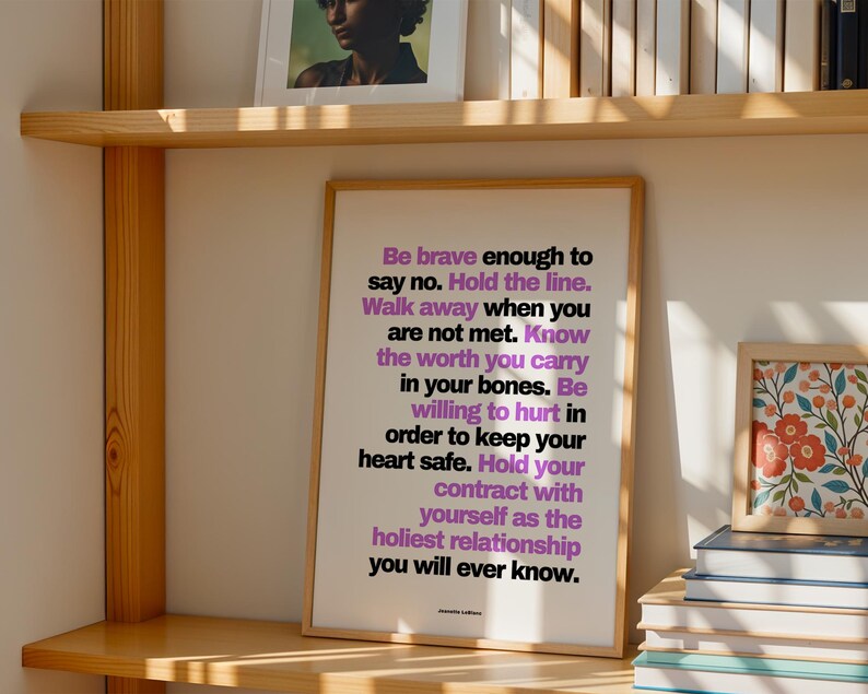 Purple and black boundary print in a light wood frame leaning on a light wood bookshelf, beside a stack of books and a small framed red and blue floral painting, with a framed portrait and a row of books on the shelf above. Warm side light. A bookish wall art moment for a library corner, and a strong woman quote to keep among your favorite books and the authors who taught you to stand firm.