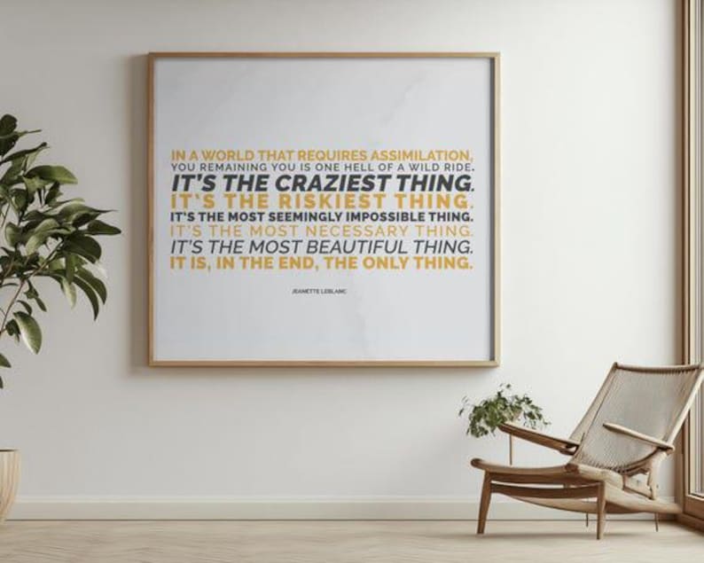 You Remaining You printable shown in a thin light wood frame hanging on a white wall in a bright, airy room, above a pale floor and beside a wood and woven lounge chair with a small plant, with a leafy branch on the left and a tall window on the right. Minimalist decor for a calm living room. A gallery wall print with inner strength in every line, from you remaining you is one hell of a wild ride to the final words.