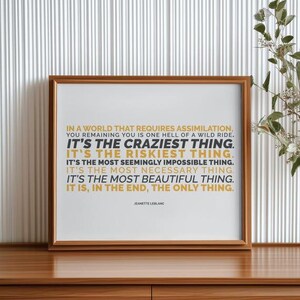 You Remaining You printable shown in a polished wood frame standing on a wood credenza against a white fluted wall panel, with green leafy stems and small white flowers to the right. Clean, modern styling for the gold and black capital lines and the name Jeanette LeBlanc. A quote printable for a living room, an entry console, or an office, with a clear message about choosing yourself in a world that asks you to conform.