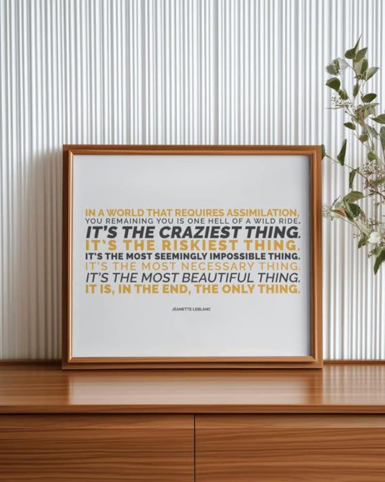 You Remaining You printable shown in a polished wood frame standing on a wood credenza against a white fluted wall panel, with green leafy stems and small white flowers to the right. Clean, modern styling for the gold and black capital lines and the name Jeanette LeBlanc. A quote printable for a living room, an entry console, or an office, with a clear message about choosing yourself in a world that asks you to conform.