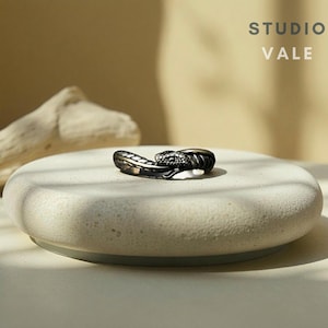 May include: A dark metallic ring shaped like a coiled snake with textured scales and a defined head. The text STUDIO VALE is visible in the upper right corner. The ring features a polished finish and a wrap-around design that forms a circular band.
