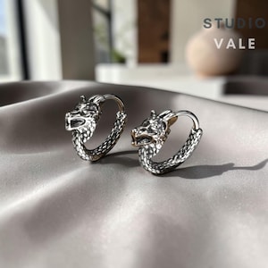 Puede incluir: Un par de pendientes de aro de plata con un diseño detallado de cabeza de lobo en la parte superior de cada aro. La parte inferior de los aros tiene un patrón texturizado similar a escamas. El texto STUDIO VALE es visible arriba a la derecha.