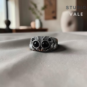 May include: A silver-toned ring featuring a detailed, textured spider design with eight prominent, dark, circular eyes. The band is crafted with intricate, hair-like engravings across the surface, and the text STUDIO VALE is visible in the upper right corner.