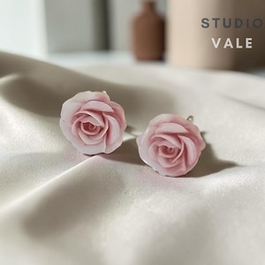 Puede incluir: Un par de pendientes de botón con forma de rosas de color rosa pálido con pétalos en capas. El texto STUDIO VALE es visible en la esquina superior derecha. Los pendientes presentan un diseño floral realista con un tono rosa claro y acabado liso.