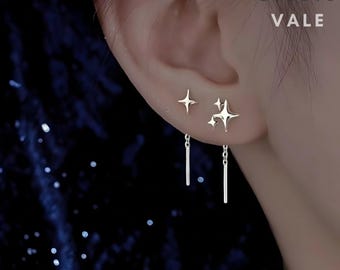 May include: Silver-toned earrings featuring four-pointed star shapes and dangling thin metal bars. One earring has a single star with a chain, while the other displays a cluster of three stars with a chain. Text reads STUDIO VALE.