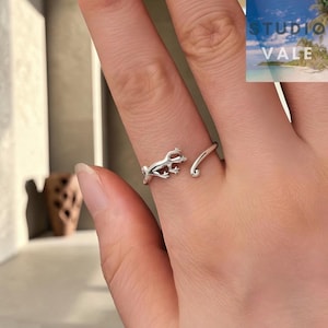 Può includere: Un anello aperto color argento con un design a lucertola scolpita. La fascia è regolabile, con un'estremità a forma di testa e corpo di lucertola e l'altra estremità che forma una semplice coda curva, creando un effetto avvolgente sul dito.
