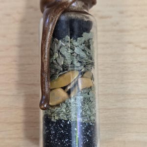 May include: A small clear glass vial sealed with a metallic bronze wax drip at the top. The bottle is filled with distinct horizontal layers of materials, including black sand, dried green herbs, smooth tan pebbles, and a dark charcoal substance.