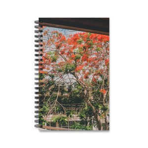 May include: A spiral-bound notebook featuring a cover image of a tree with vibrant orange-red flowers and green leaves, partially obscured by dark horizontal lines and a dark brown architectural element at the top of the frame.
