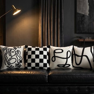 Peut inclure: Quatre coussins carrés disposés en rangée. Le premier présente un visage dessiné au trait noir, le deuxième un motif à damier noir et blanc, le troisième un gribouillis noir continu et le quatrième une série de lignes noires épaisses.