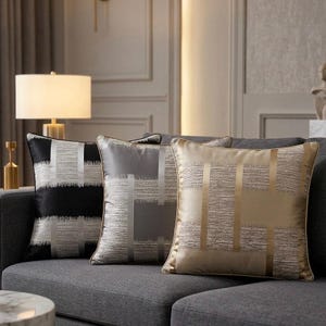 Peut inclure: Trois coussins carrés présentant un motif géométrique composé de bandes verticales et horizontales unies entrecoupées de panneaux texturés aux bords effilochés. Les coussins sont noirs, gris argenté et or métallique, avec un passepoil assorti.