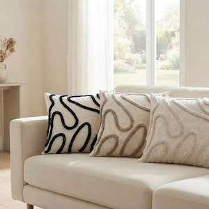 Peut inclure: Trois coussins carrés disposés sur un canapé de couleur claire. Le coussin de gauche est blanc cassé avec des lignes abstraites, ondulées et touffetées noires. Les deux autres sont blanc cassé avec des lignes touffetées assorties.