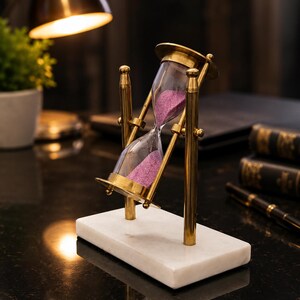 May include: A rotating hourglass featuring a clear glass bulb filled with purple sand, supported by a polished brass frame. The hourglass is mounted on a rectangular white marble base, allowing the glass component to pivot between two vertical brass pillars.