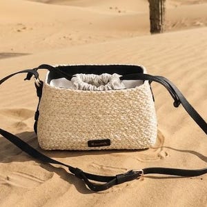 Puede incluir: Un bolso tipo cubo de paja trenzada color crema con una correa de hombro negra ajustable y un cierre de cordón con interior de tela beige claro. Una pequeña etiqueta rectangular negra con texto en blanco está fijada en la parte frontal.