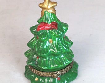 May include: A glossy green ceramic Christmas tree figurine with a gold-toned star topper, a red bow detail, and small gold and red accents. The tree features a hinged metal band around the middle, allowing the top section to open as a small container.